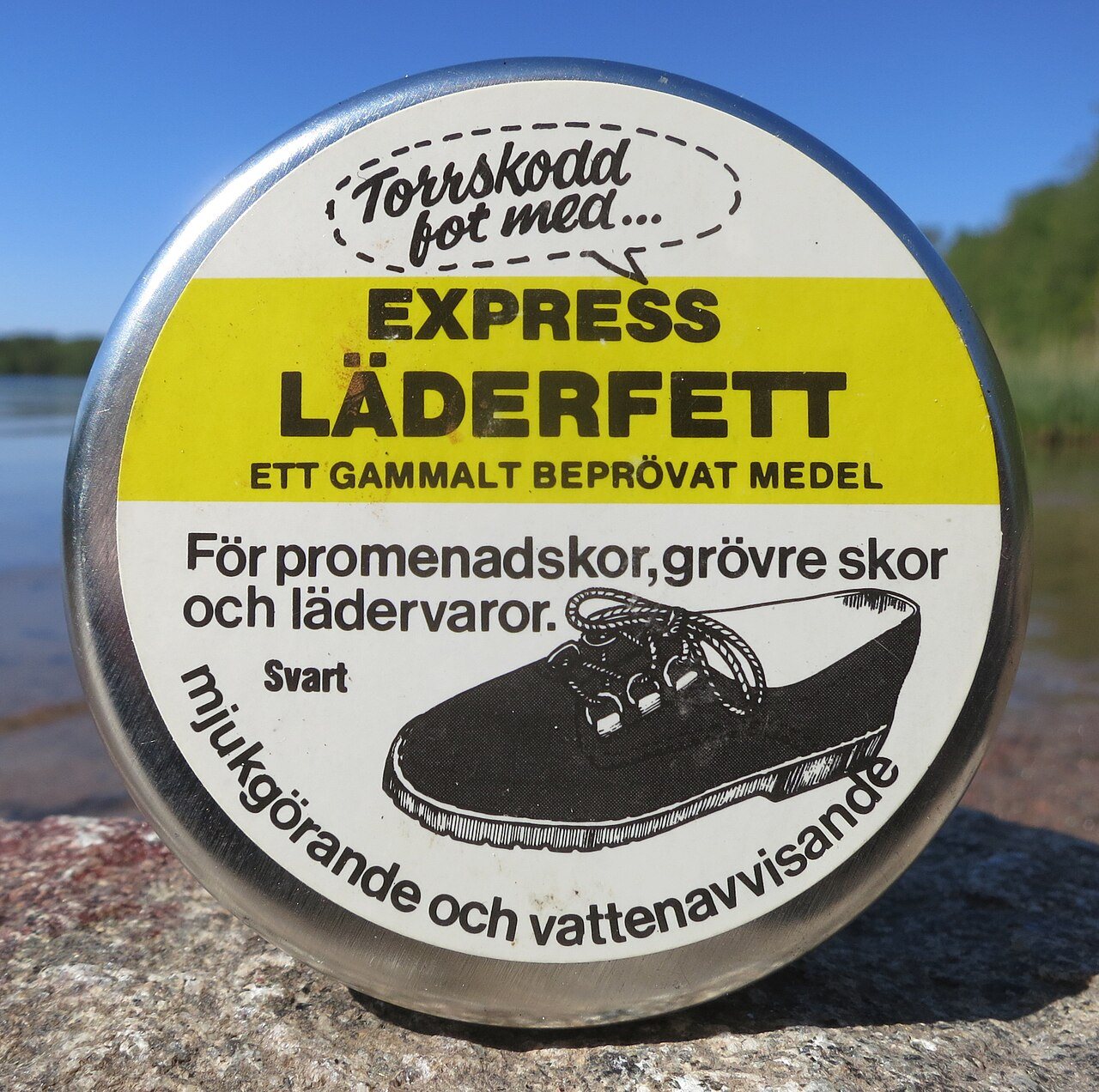 A round 1990s Swedish tin of black leather grease for walking shoes, with a drawing of a lace-up shoe on the lid, standing on a rock by a lake